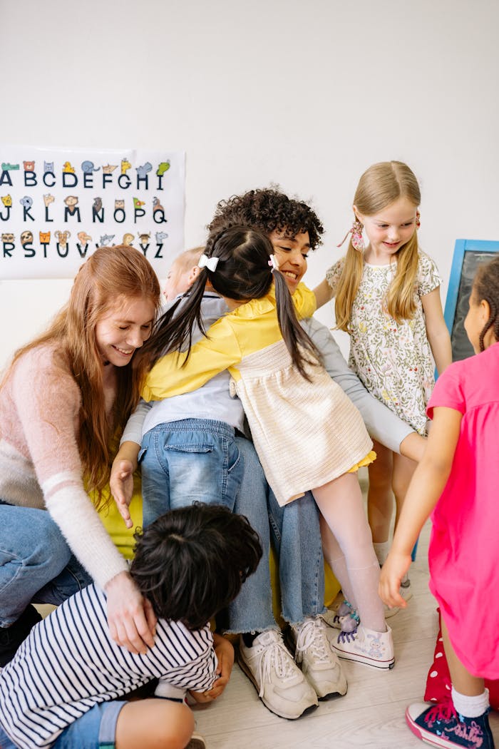 A group of diverse preschool children warmly hug their smiling teacher in a colorful classroom setting.